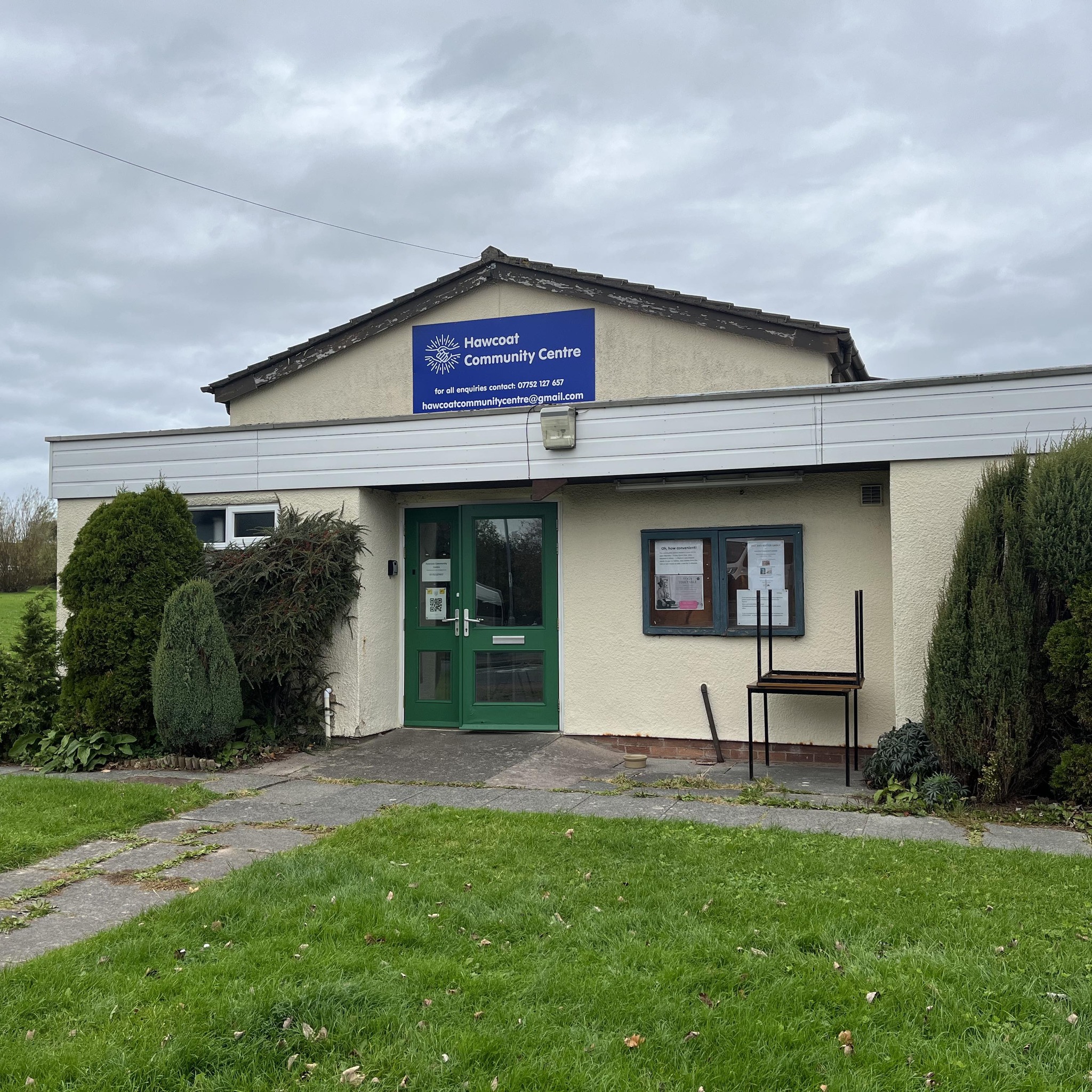 A cream painted single story building with a pithed tiled roof. There are green glazed double doors and a noticeboard to the front, with bushes in front of a window. A large blue sign has the Centre's name on.