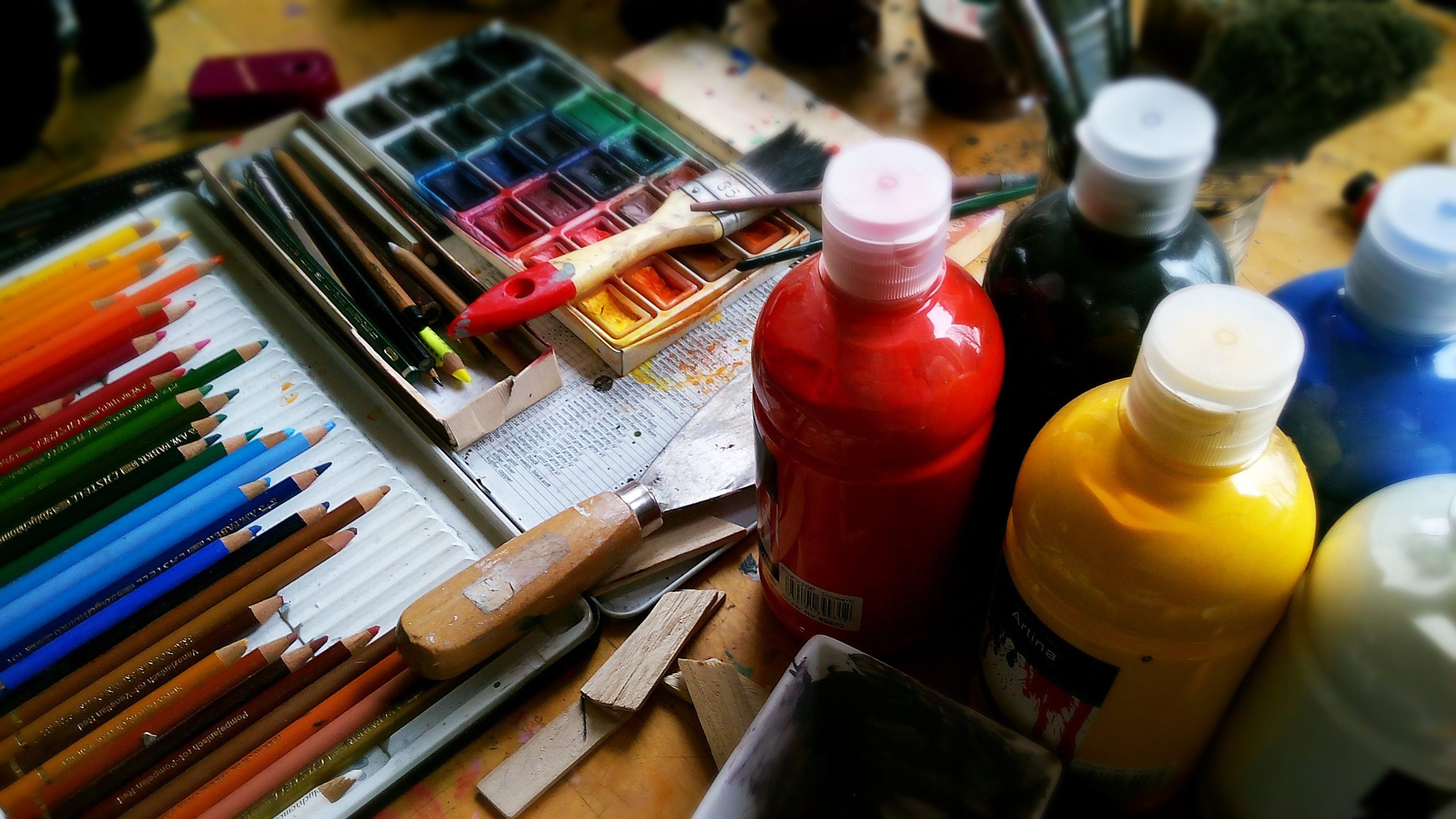 Photo of art materials, including bottles of paint in bright colours, pencils grouped together by colour, paintbrushes and a water-colour paintbox