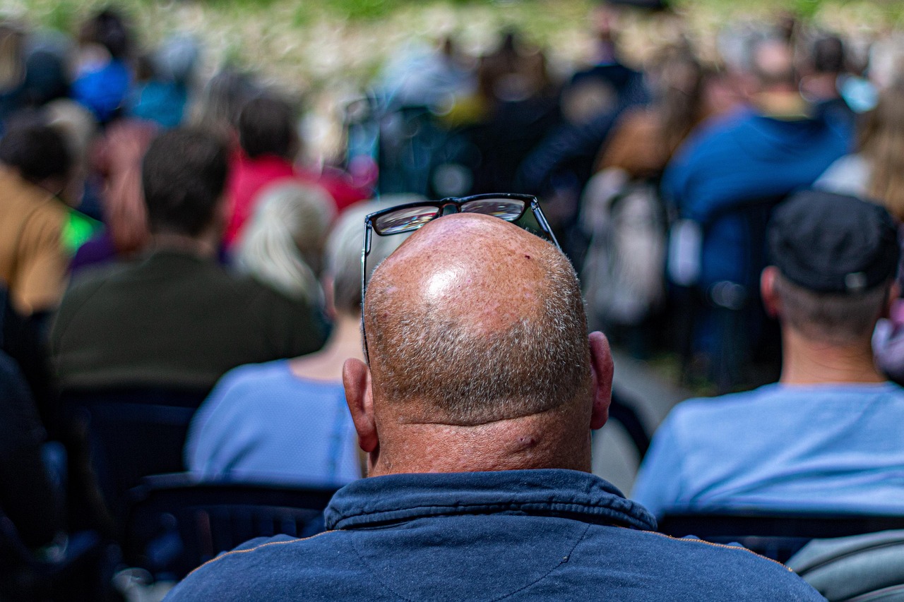 A photo showing the back of a balding man's head, with glasses propped on his forehead. He is wearing a blue fleece. Other people are sat around and in front of him, but blurred.