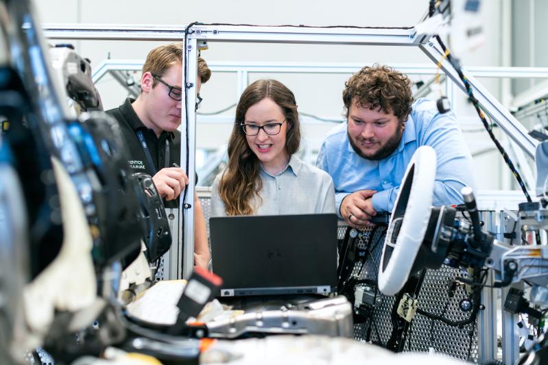 Three students (2 male and one female) are stood at workbench looking at a laptop. Various items of electrical equipment cover the table in front of them. They are smiling. All have brown hair.