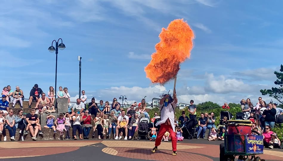A photo of a man dressed in white lab coat a red trousers, with a large ball of flame above him. A crowd, in an outdoor paved space, watches on.