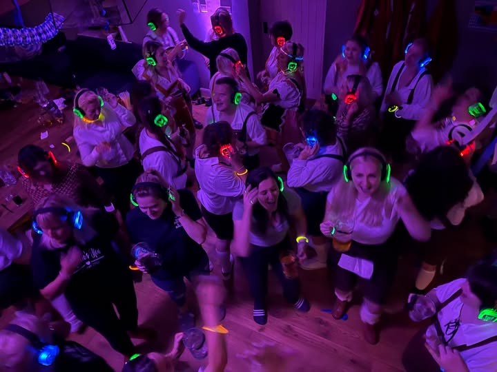 A photo of people bathed in purple light on a dancefloor. They are dancing a smiling, all wearing headphones which glow green, red or blue.