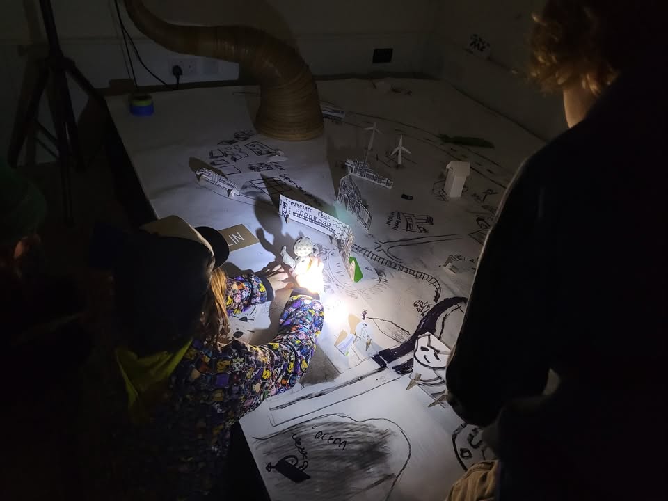 A child is holding a model, on a table covered in drawings and drawings turned in to stand-up pieces. Its dark, but what the child is holding is illuminated by a torch.