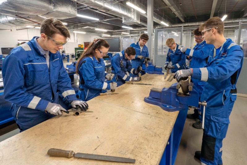 Young people wearing blue overalls and protective goggles a working on pieces of pipe around a wooden bench.